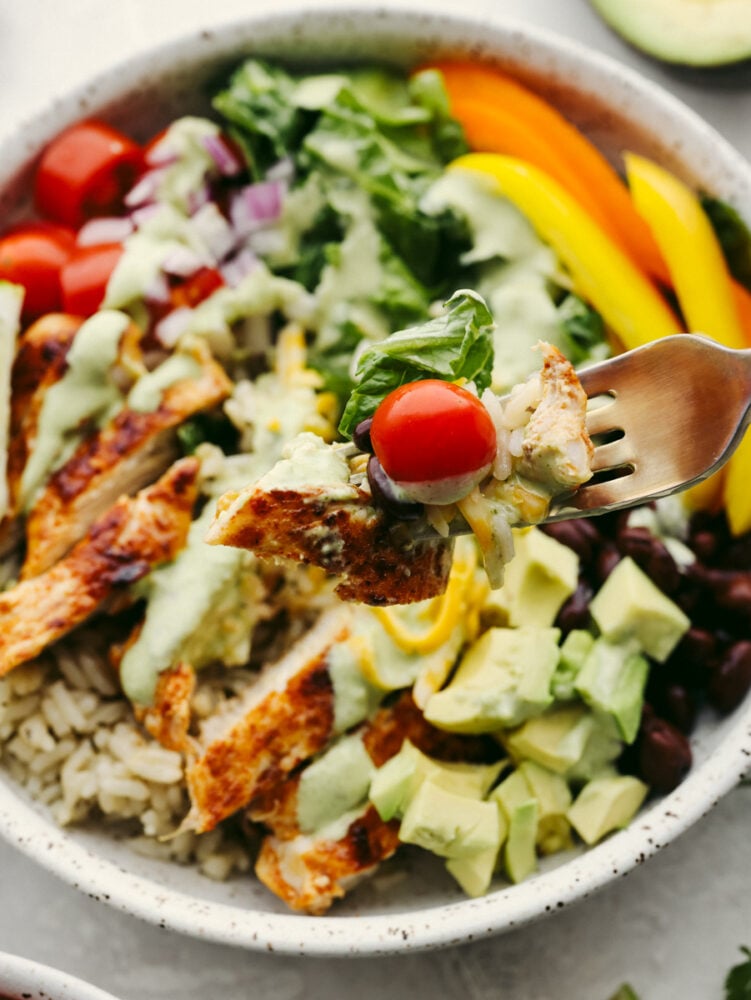 Packed with fresh veggies, rice, and juicy seared chicken, burrito bowls are the most delicious way to fuel your body! Topped off with creamy cilantro ranch, this is the perfect lunch filled with amazing flavor and texture. Taking a bite of chicken and vegetables with a fork.