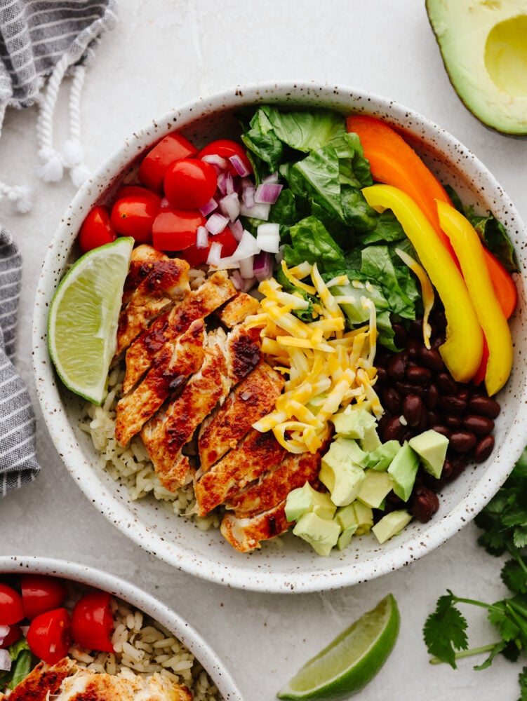 Packed with fresh veggies, rice, and juicy seared chicken, burrito bowls are the most delicious way to fuel your body! Topped off with creamy cilantro ranch, this is the perfect lunch filled with amazing flavor and texture. Chicken, vegetables, and beans in a white stoneware bowl.