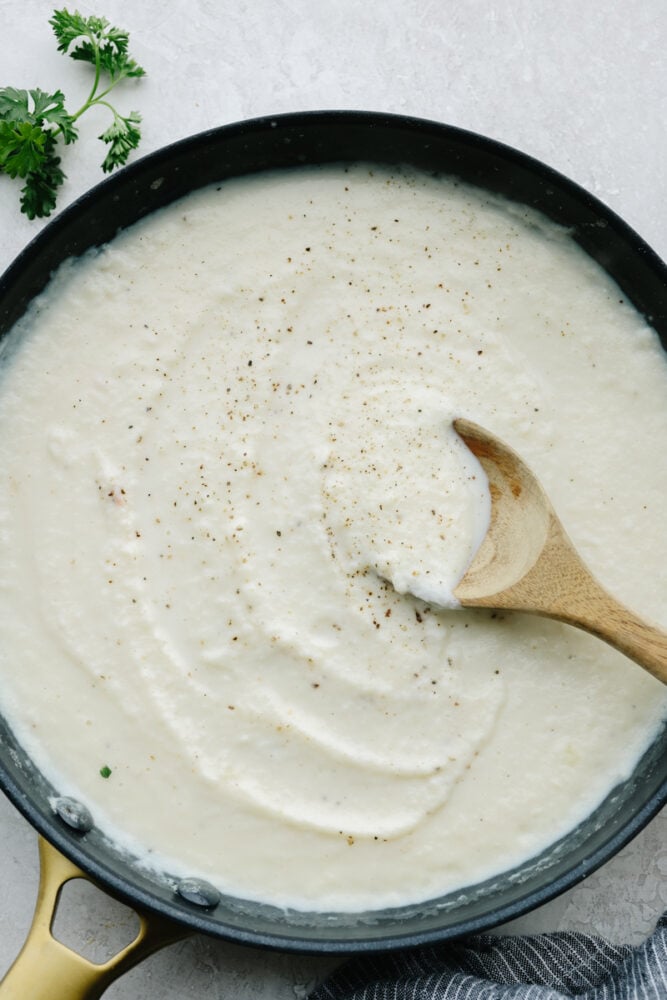 This cauliflower alfredo sauce is rich, creamy, and healthy! With this sauce, you get all of that insanely delicious flavor without the heavy cream. It's so good that you will want to put it on everything! Cauliflower alfredo sauce in a saucepan with a wooden spoon.