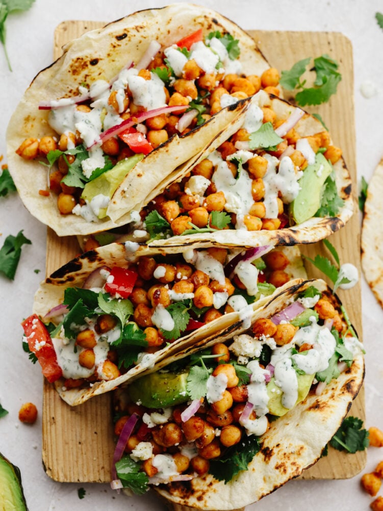 Chickpea tacos are a great way to get your daily protein and all the Tex-Mex flavors that you crave! These flavorful tacos are loaded with fresh veggies and topped off with a creamy cilantro drizzle! Closeup of 4 chickpea tacos laid out on a wooden board.