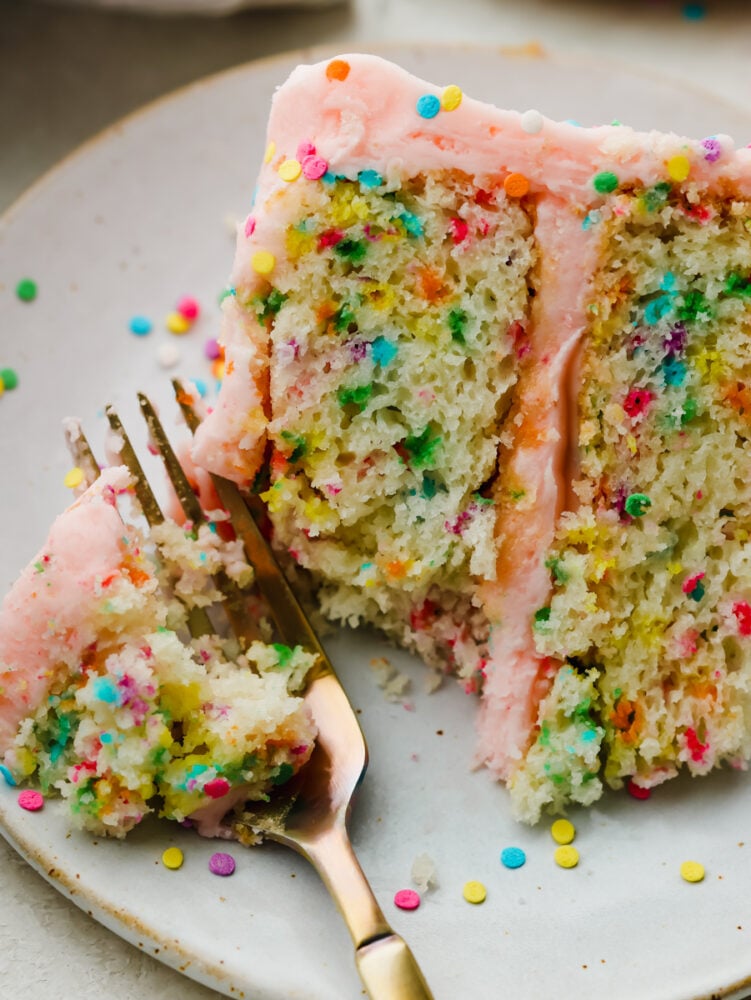 This light and fluffy homemade funfetti cake is perfect for every celebration! A white cake bursting with rainbow sprinkles is so delicious and will have everyone asking for a second slice. A slice of funfetti cake on a plate being cut with a gold fork.