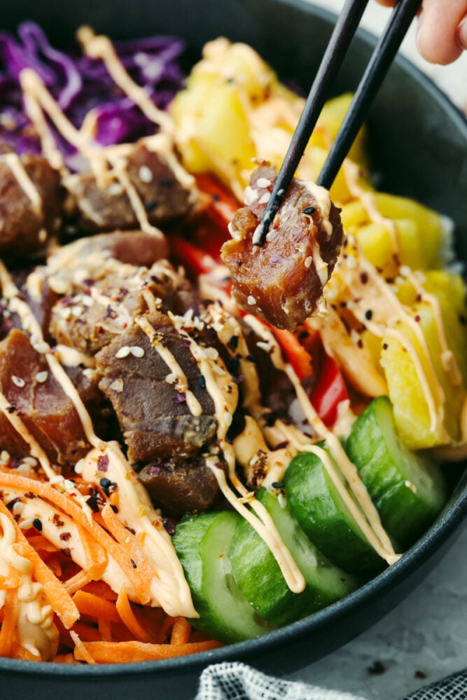 This Hawaiian poke bowl is fantastically fresh and full of healthy ingredients! Skip ordering this at a restaurant and make it at home in less than 15 minutes!ย A hand holding chopsticks with a piece of tuna coming out of a Hawaiian poke bowl