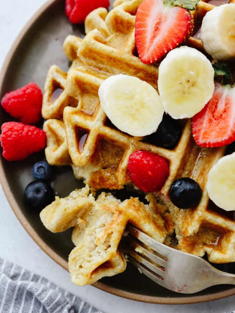 Get ready for some delicious and healthy waffles that are going to knock your socks off! These protein waffles are absolutely delicious, and you can whip them up in less than 10 minutes! Zoomed in shot of a waffler on a plate. It's topped with fruit and being cut into with a fork.