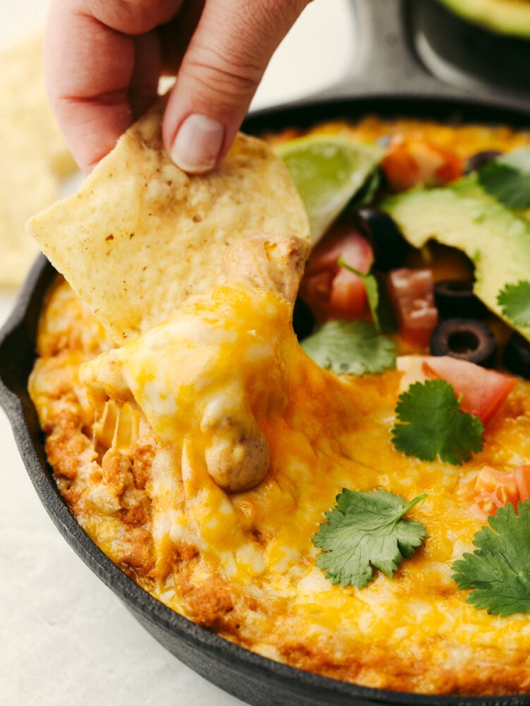 Texas trash dip is a classic warm bean dip that gets loaded with corn, olives, black beans, and ooey gooey cheese! This dip will be the hit at your next party!ย A tortilla chip being dipped into Texas trash dip.