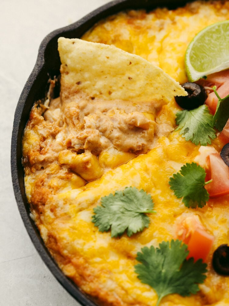 Texas trash dip is a classic warm bean dip that gets loaded with corn, olives, black beans, and ooey gooey cheese! This dip will be the hit at your next party!ย A close up shot of a tortilla chip in a pan of hot bean dip.