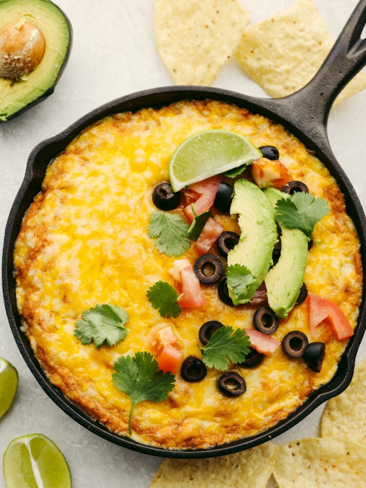 Loaded Texas Trash dip is the classic warm bean dip that gets loaded with corn, olives, black beans, and ooey gooey cheese! This dip will be the hit at your next party!ย A pan of Texas trash dip garnished with olives, cilantro, a lime wedges and some tomatoes and avocado.