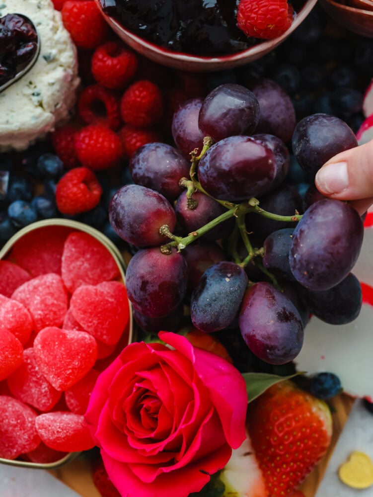 The holiday of love is just around the corner, and you need to try out this Valentine's day charcuterie board! It's packed with meat, cheeses and all sorts of festive red and pink fruits and candy to start the holiday off right. A close up of a hand picking some grapes off of the board.