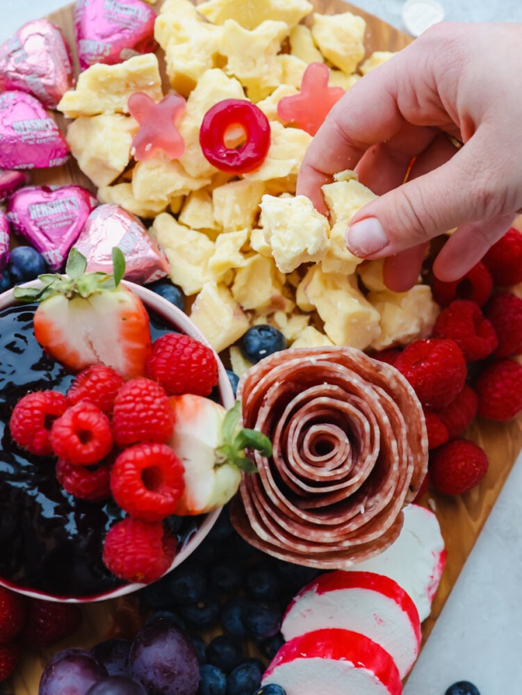 The holiday of love is just around the corner, and you need to try out this Valentine's day charcuterie board! It's packed with meat, cheeses and all sorts of festive red and pink fruits and candy to start the holiday off right. A close up shot of a salami rose and someone picking up a piece of cheese.
