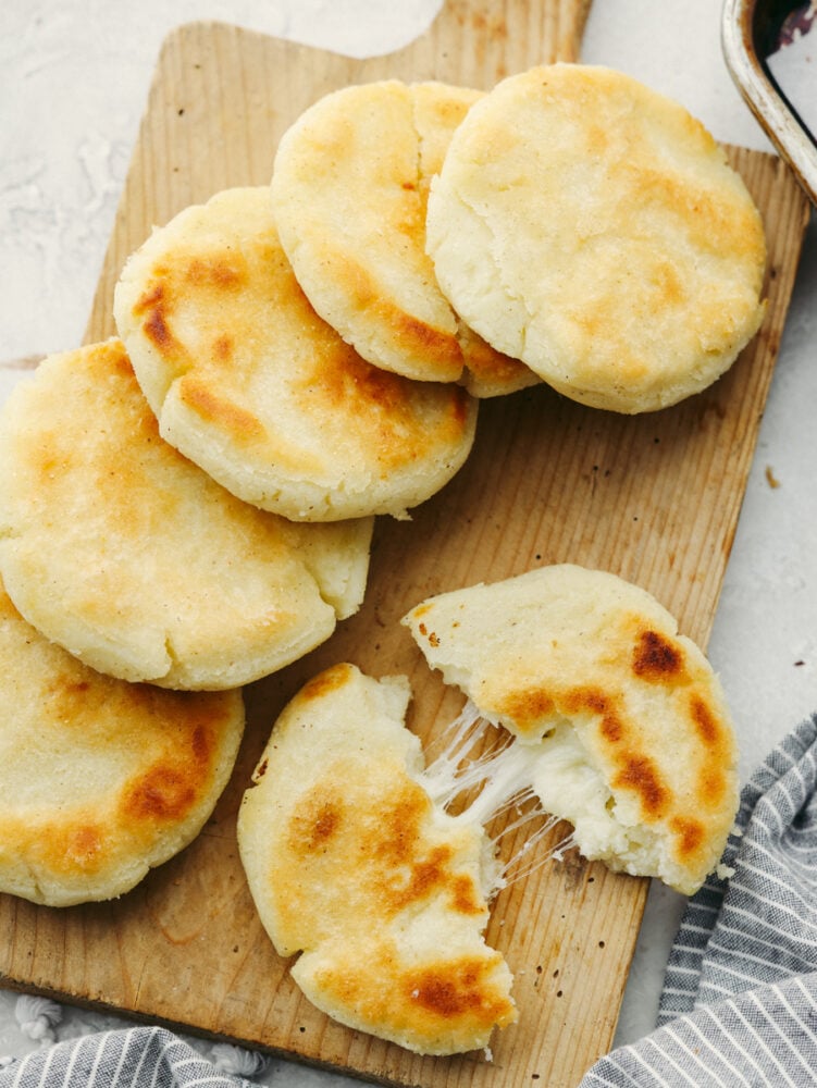 Arepas are a delicious cheesy bread made out of cornmeal! This super simple Colombian treat is sure to win the hearts of your family. Arepas on a wooden serving platter with one ripped in half.