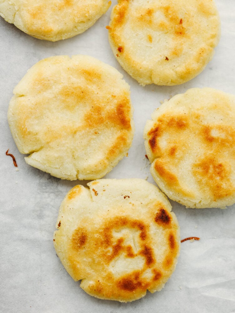 Arepas are a delicious cheesy bread made out of cornmeal! This super simple Colombian treat is sure to win the hearts of your family. Disc-shaped breads on a baking sheet.