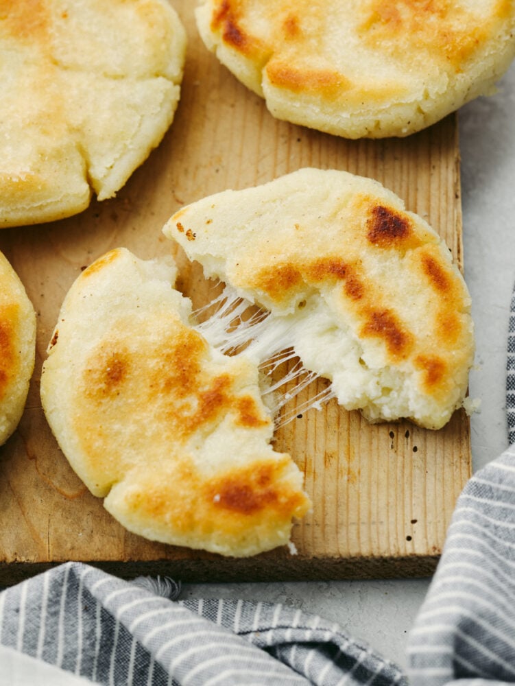 Arepas are a delicious cheesy bread made out of cornmeal! This super simple Colombian treat is sure to win the hearts of your family. An arepa broken in half on a wooden board.