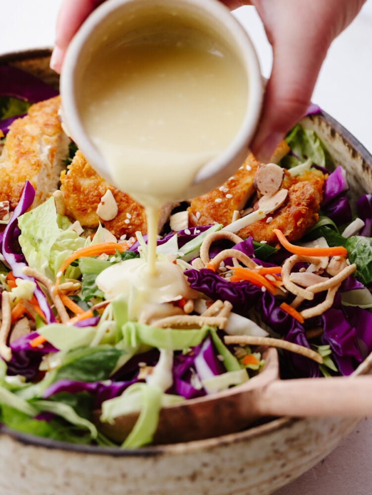 This Asian chicken salad is packed with crispy greens, crunchy chicken, toasted almonds, and chow mein noodles. It's tossed with a light and delicious creamy vinaigrette and will be your new go-to salad! A bowl of asian chicken salad with a creamy vinaigrette being poured on top.