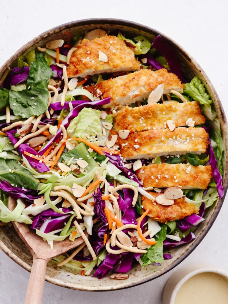 This Asian chicken salad is packed with crispy greens, crunchy chicken, toasted almonds, and chow mein noodles. It's tossed with a light and delicious creamy vinaigrette and will be your new go-to salad! A top view of salad greens with a sliced chicken breast on top.