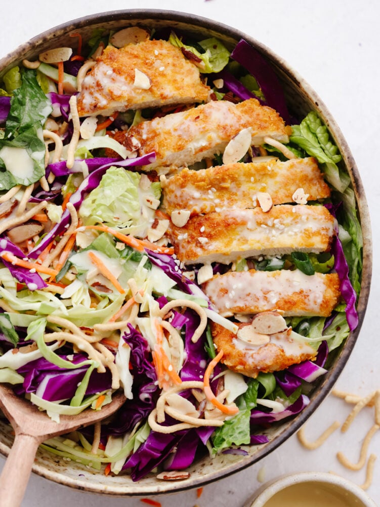 This Asian chicken salad is packed with crispy greens, crunchy chicken, toasted almonds, and chow mein noodles. It's tossed with a light and delicious creamy vinaigrette and will be your new go-to salad! The top view of Asian chicken salad with dressing in a bowl with a wooden spoon.