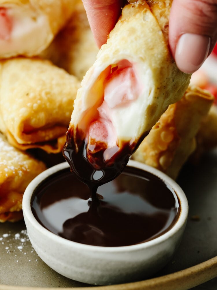 Crispy on the outside with a creamy, strawberry filling on the inside, these cheesecake egg rolls are the BEST. Your guests will eat one and won't be able to stop! A cheesecake egg roll being dipped in chocolate sauce.