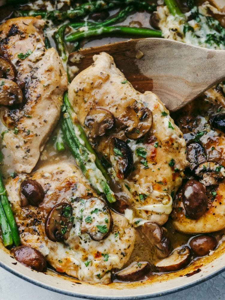 Juicy chicken is covered in creamy Madeira sauce, melty cheese, mushrooms, and asparagus in this easy skillet recipe! Chicken Madeira brings all of those delicious Cheesecake Factory flavors right to your kitchen! Closeup of scooping up a piece of chicken Madeira with a wooden spoon.