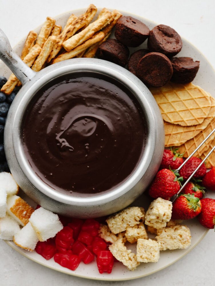 The best way to impress your valentine is with homemade chocolate fondue! This silky-smooth dessert is so amazing and pairs perfectly with all of your favorite treats! A pot of fondue surrounded by fruit, cookies, and treats.