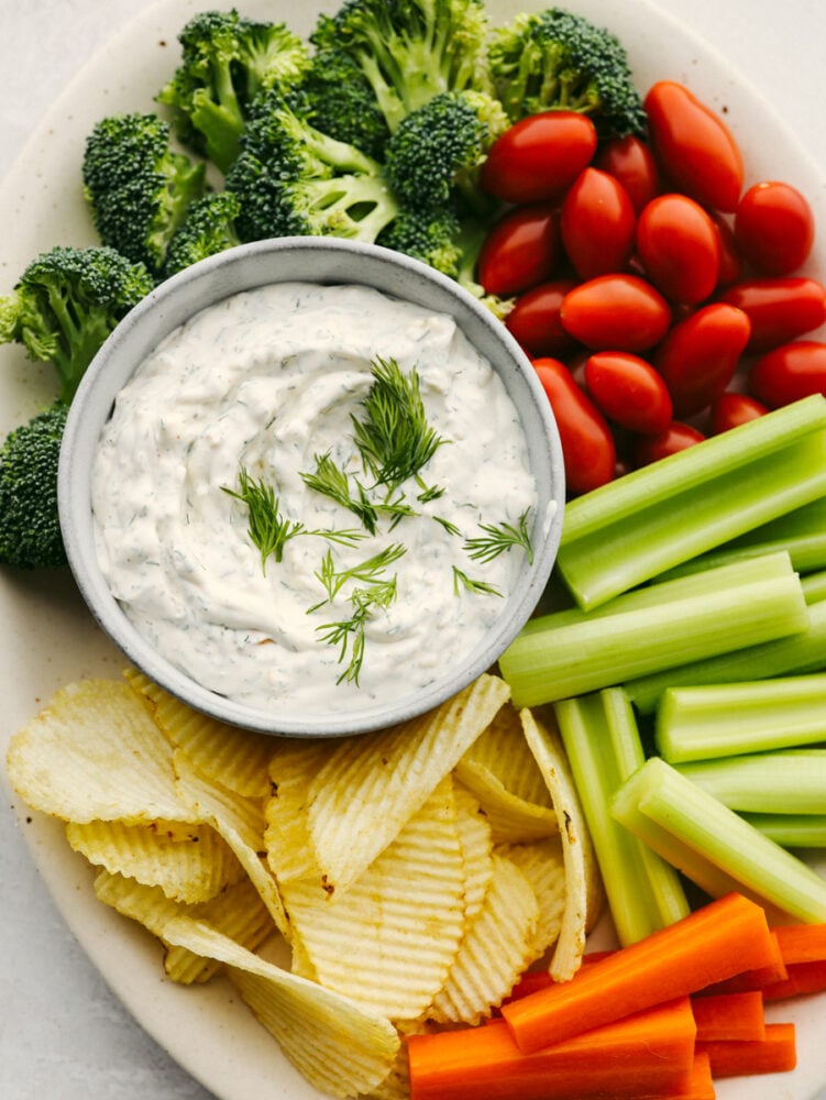This delicious dill dip is easy and goes with everything. It is creamy and full of flavor and will be ready to eat in less than 5 minutes! If you are a dill pickle lover like me then you are going to be bringing this to every party and get-together! A plater with dill dip in a bowl surrounded by chips, celery, tomatoes and broccoli.