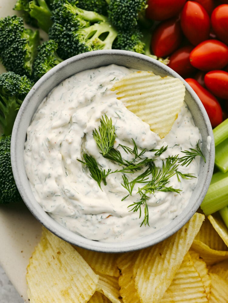 This delicious dill dip is easy and goes with everything. It is creamy and full of flavor and will be ready to eat in less than 5 minutes! If you are a dill pickle lover like me then you are going to be bringing this to every party and get-together! A top view of some dill dip on a platter with chips, broccoli, celery and cherry tomatoes.