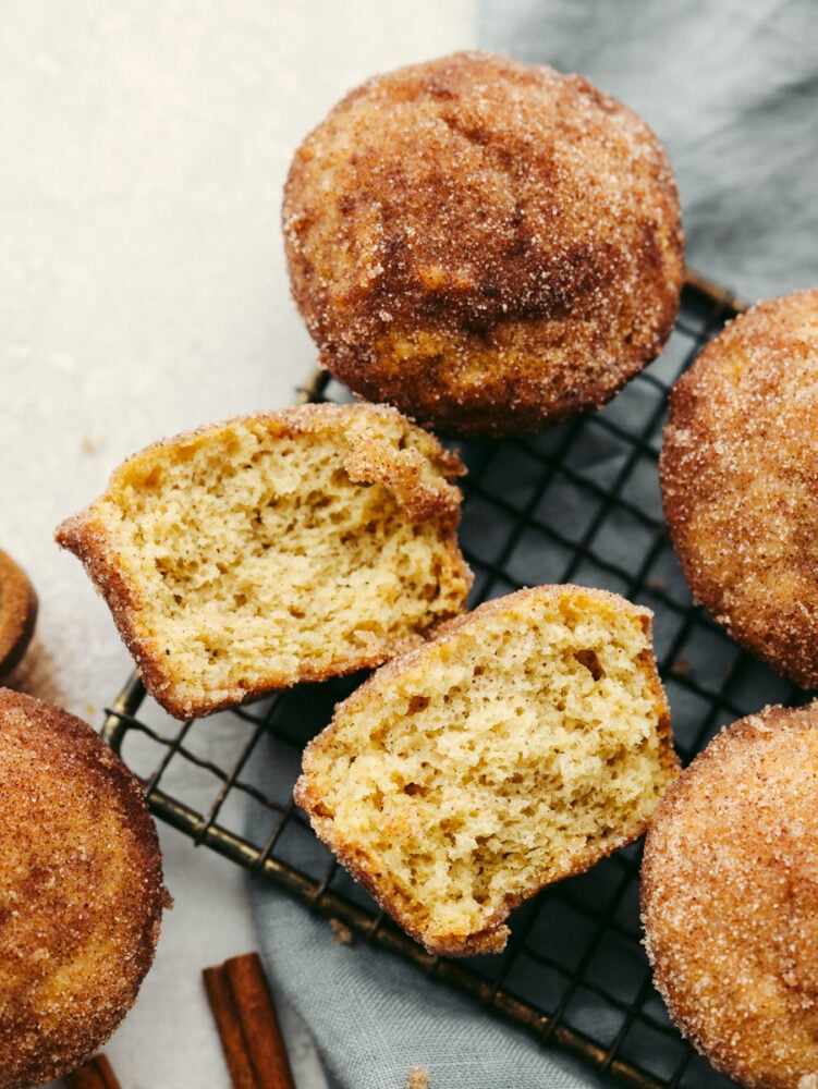 Donut muffins are soft and fluffy, with a cinnamon-sugar flavored goodness that melts in your mouth! They are delicious enough for anyone to want to devour them the moment that you put them on the table! Donut muffins on a cooling rack with one broken in half so that you can see the inside.
