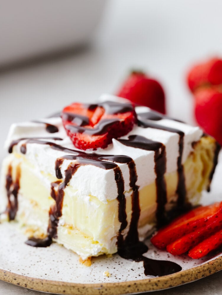 Eclair cake is moist and sweet, made with vanilla filling, a buttery crust, and whipped cream topping. There are so many toppings to choose from and you will be amazed at how insanely soft and delicious this cake is! A slice of eclair cake topped with chocolate drizzle and sliced strawberries.