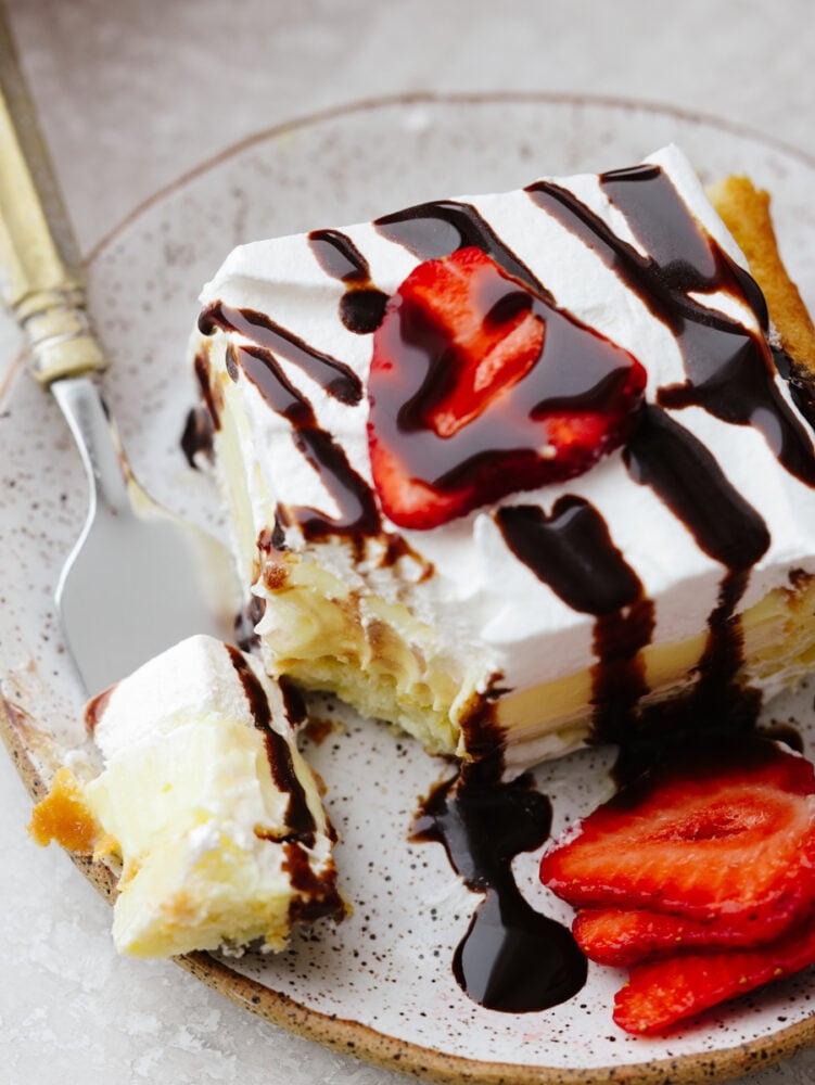 Eclair cake is moist and sweet, made with vanilla filling, a buttery crust, and whipped cream topping. There are so many toppings to choose from and you will be amazed at how insanely soft and delicious this cake is! Taking a bite of eclair cake topped with strawberries and chocolate sauce.