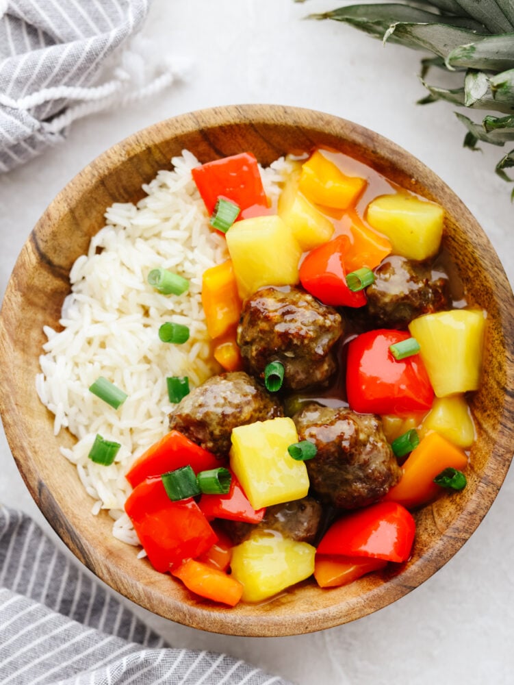 Hawaiian meatballs are the PERFECT main course for your dinner! You don't want to miss out on these! They are drenched in the most delicious sauce and packed with flavor. A wooden bowl filled with rice, veggies and meatballs. All garnished with green onions.