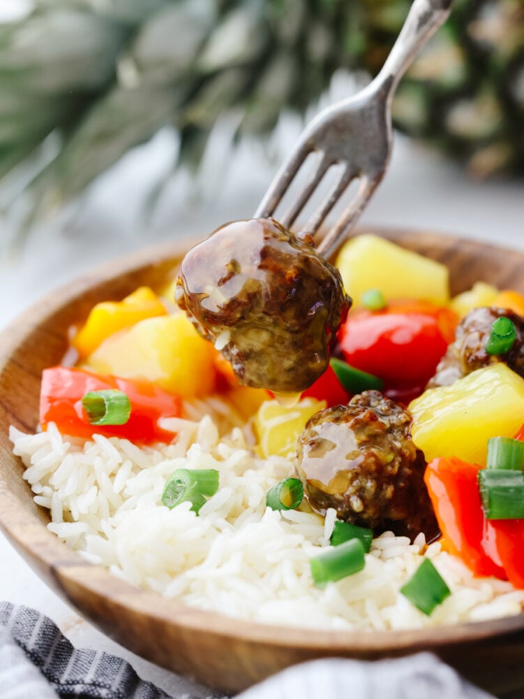 Hawaiian meatballs are the PERFECT main course for your dinner! You don't want to miss out on these! They are drenched in the most delicious sauce and packed with flavor. A wooden bowl filled with rice, meatballs and vegetables. One meatball is being poked with a fork.