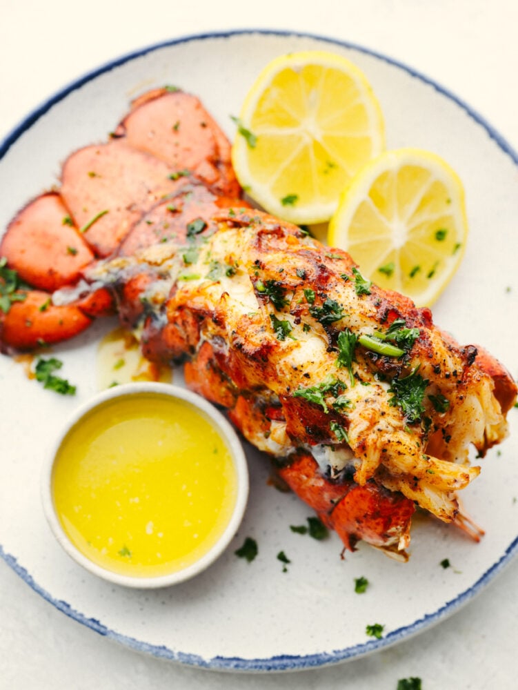 These lobster tails cooked in the air fryer are incredibly tender and delicious. Topped with a melted buttery garlic herb sauce, this is a quick and easy way to have that delectable fancy dinner without a lot of time and preparation! Lobster on a plate with lemon slices and butter.