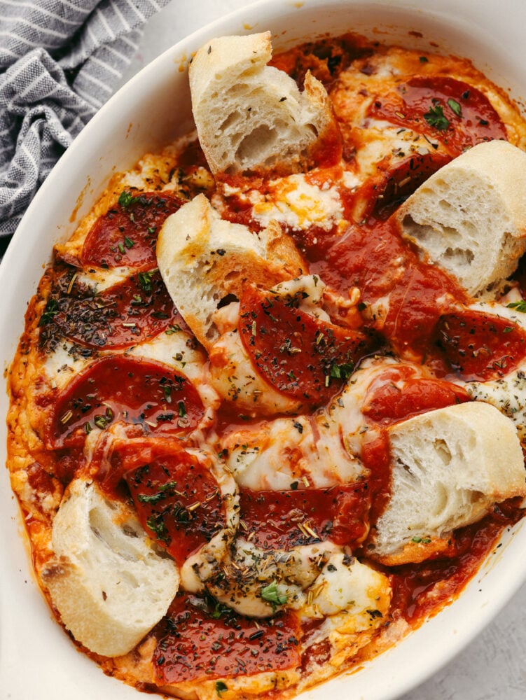 Pepperoni pizza dip is ooey-gooey cheese mixed with flavorful pizza seasonings and topped with delicious pepperoni. It is baked until it's hot and bubbly, and it will be the perfect appetizer at any party! Bread being dipped into pepperoni pizza dip.