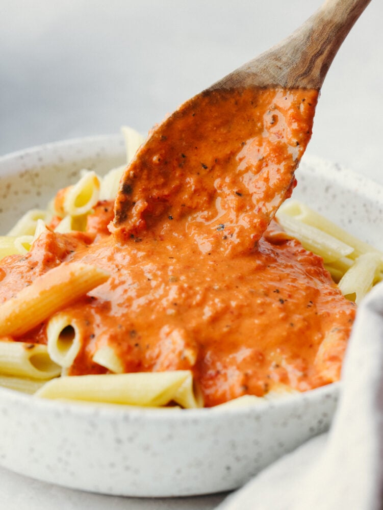 Level up pasta night with this irresistible roasted red pepper sauce! It's creamy, cheesy, and delicious with smoky, sweet spice. Be sure to make enough for seconds, your family won't be able to get enough! Pouring sauce over penne pasta with a wooden spoon.