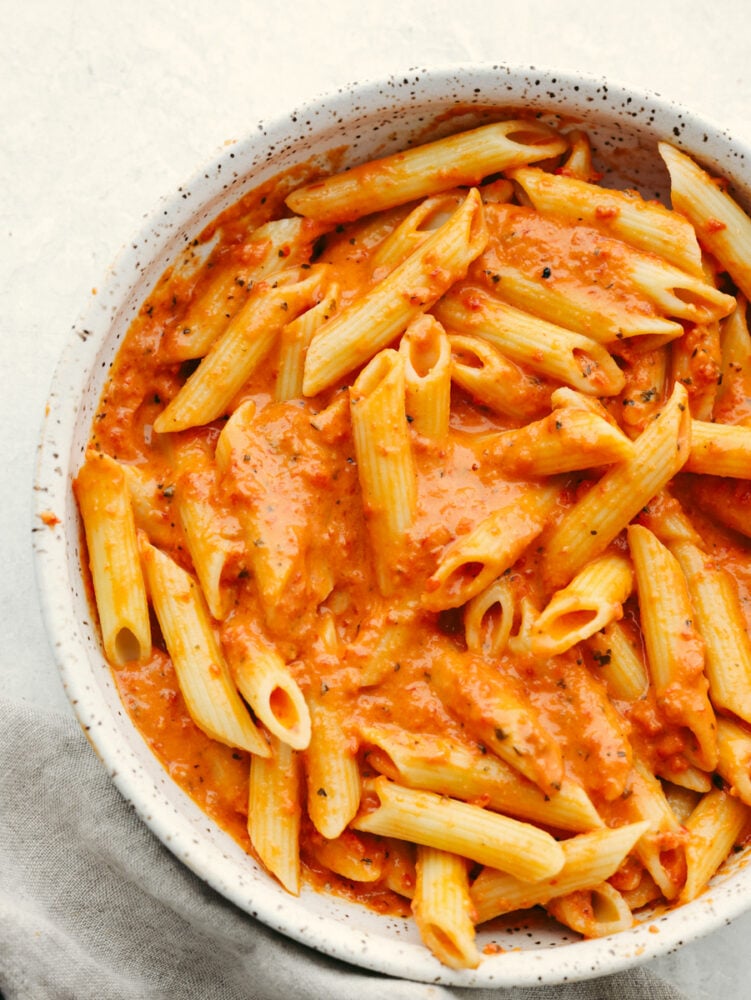 Level up pasta night with this irresistible roasted red pepper sauce! It's creamy, cheesy, and delicious with smoky, sweet spice. Be sure to make enough for seconds, your family won't be able to get enough! Penne coated with red pepper pasta sauce in a white speckled stoneware bowl.