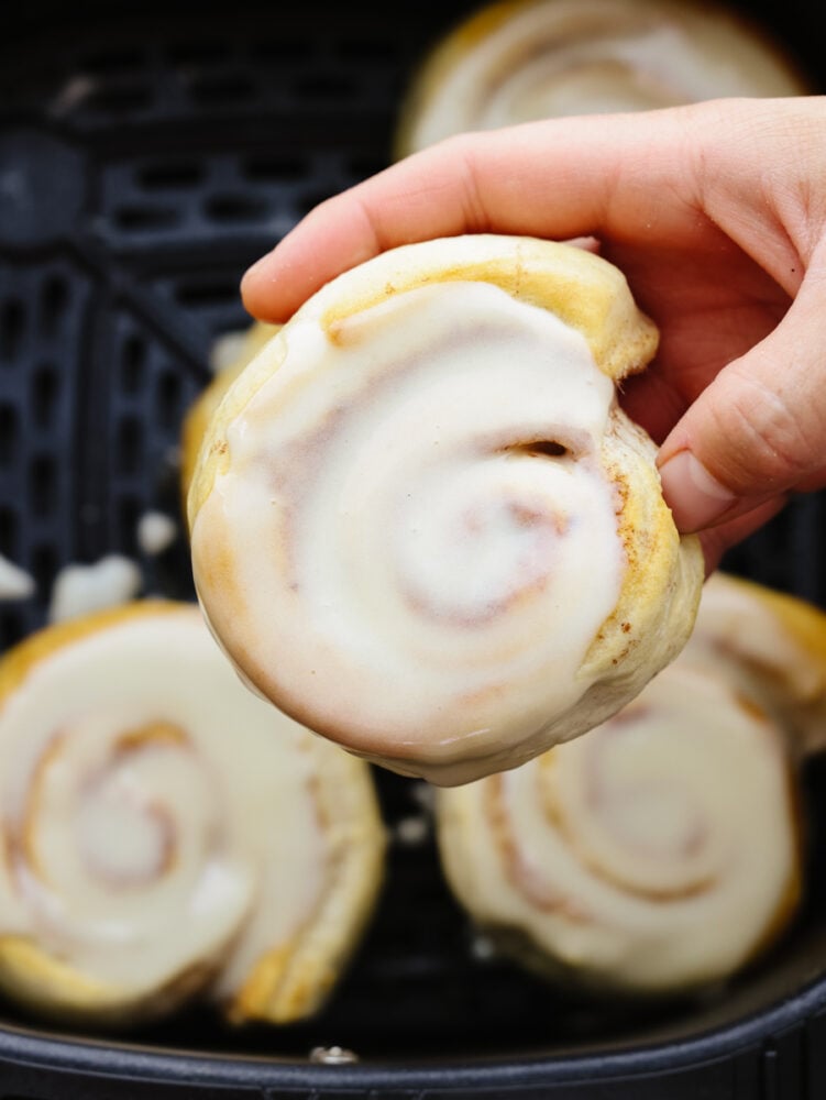 There is no easier way to cook cinnamon rolls out there! These air fryer cinnamon rolls from a can are so easy and delicious. They take less than 15 minutes to bake! A close up of someone holding a frosted cinnamon roll.