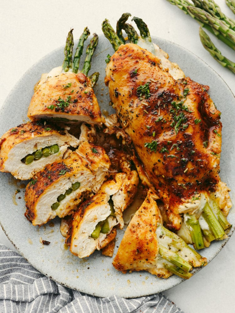 This asparagus stuffed chicken is incredibly juicy and tender. Flavored with the most delicious spices and stuffed with the incredibly healthy vegetable, asparagus, you will want to lick your plate clean! A gray plate with one whole asparagus stuffed chicken breast and another breast that is sliced into 5 pieces.