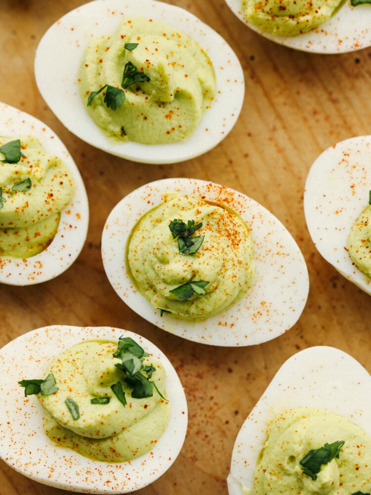 A twist on a classic appetizer, avocado deviled eggs are so creamy and delicious! Flavored with lime and dusted with paprika, your guests won't be able to get enough of this flavorful dish! Avocado deviled eggs on a wooden board, dusted with paprika.