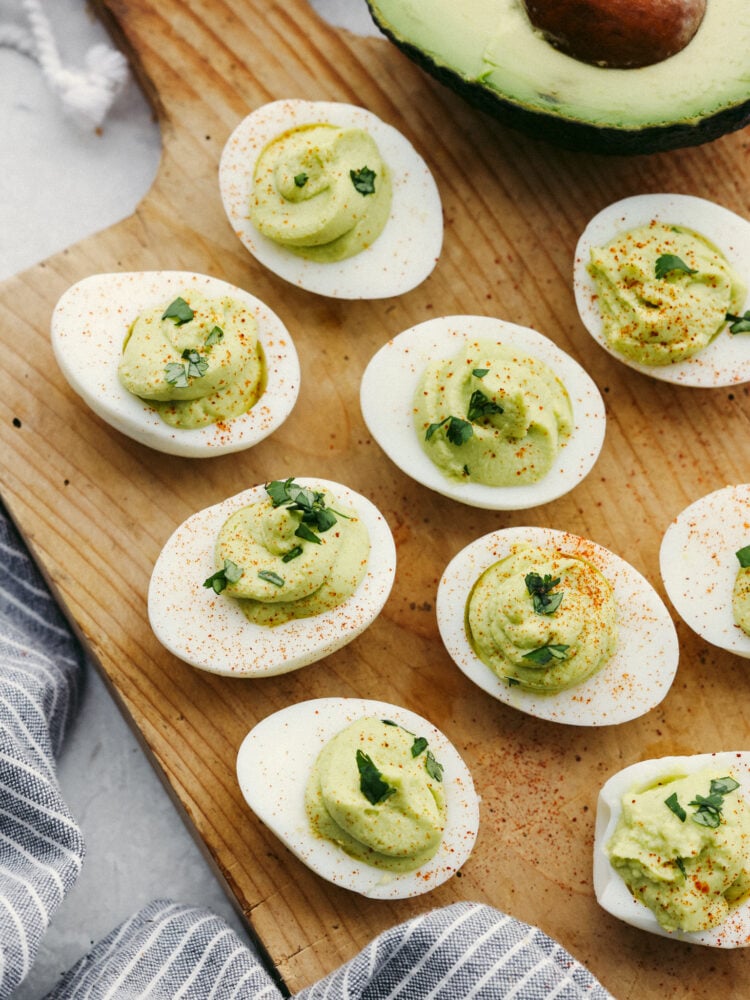 A twist on a classic appetizer, avocado deviled eggs are so creamy and delicious! Flavored with lime and dusted with paprika, your guests won't be able to get enough of this flavorful dish! Top-down view of deviled eggs on a wooden board.
