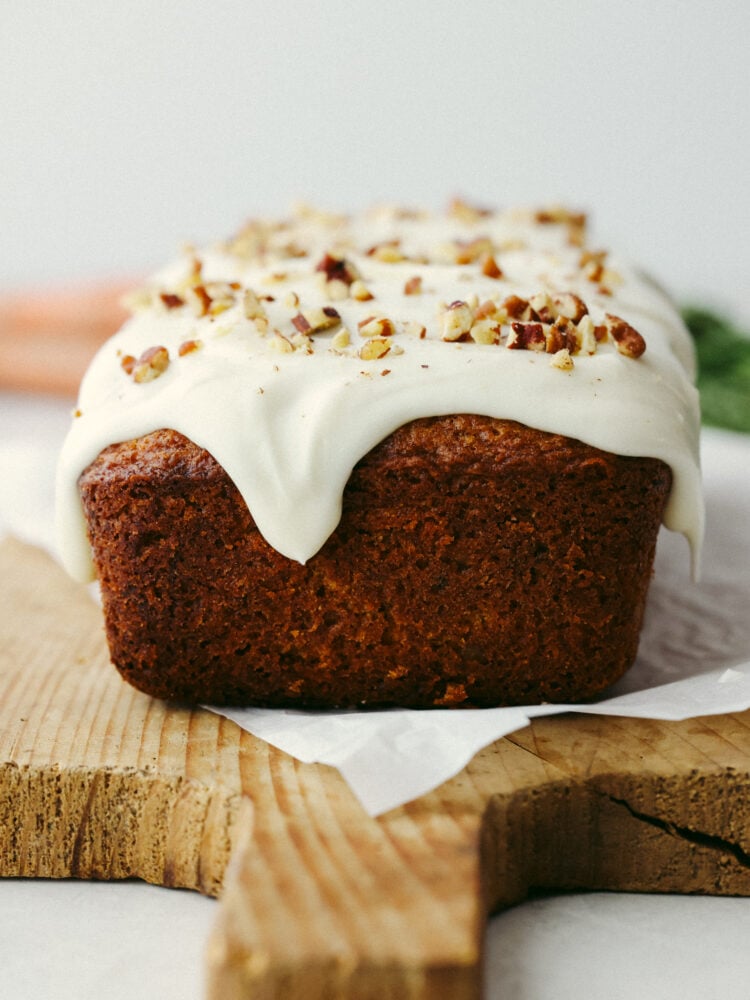 This carrot cake bread is to die for! It's moist and soft and has the most incredible flavor that tastes like cake, but it is half the work involved! This is perfect for Easter and springtime! A side view of a loaf of bread sitting on a wooden cutting baord with a glaze and nuts on top.