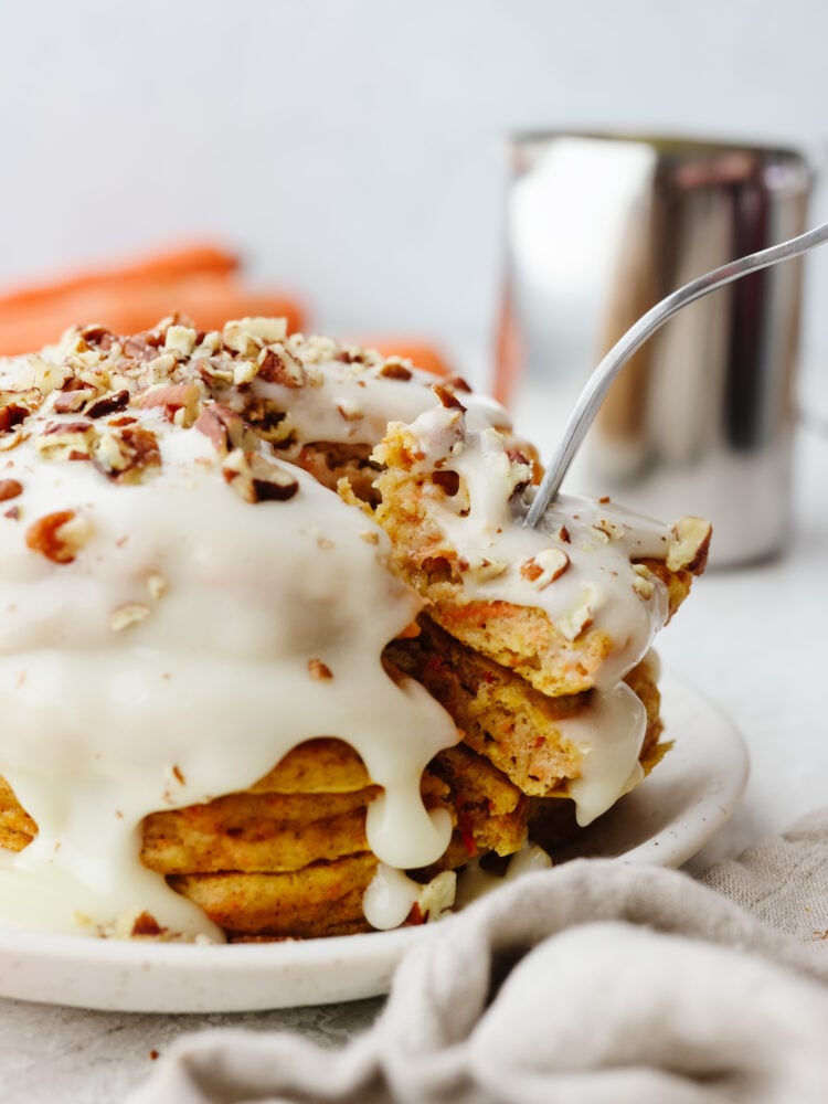 These carrot cake pancakes are fluffy, moist, and drizzled with a delicious cream cheese glaze that makes my mouth water! They will get gobbled up and fly off your table! A stack of carrot cake pancakes with a carrot cake glaze with a fork cutting into them.
