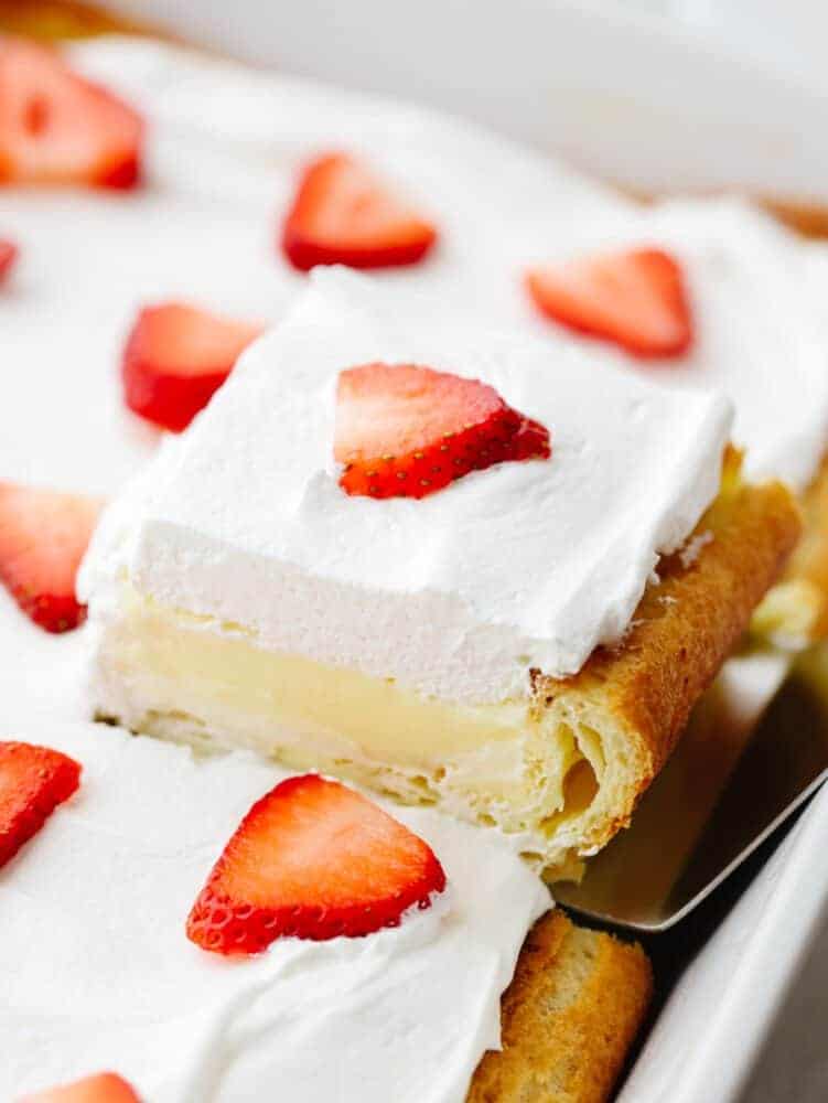 Eclair cake is moist and sweet, made with vanilla filling, a buttery crust, and whipped cream topping. There are so many toppings to choose from and you will be amazed at how insanely soft and delicious this cake is! Picking up a slice of eclair cake with a cake spatula.