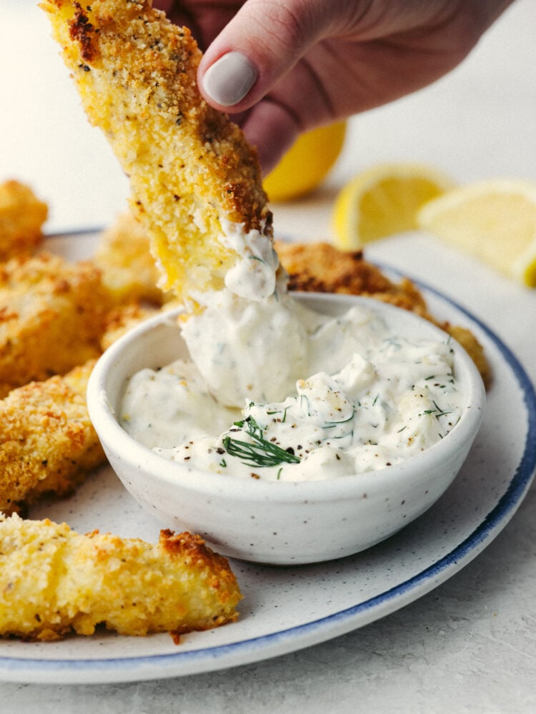 Oven-baked fish sticks are crispy, tender, and SO good! Coated with breadcrumbs and delicious seasoning, these fish sticks are packed with flavor and a recipe that the whole family will enjoy! A fish stick being dipped in tartar sauce.