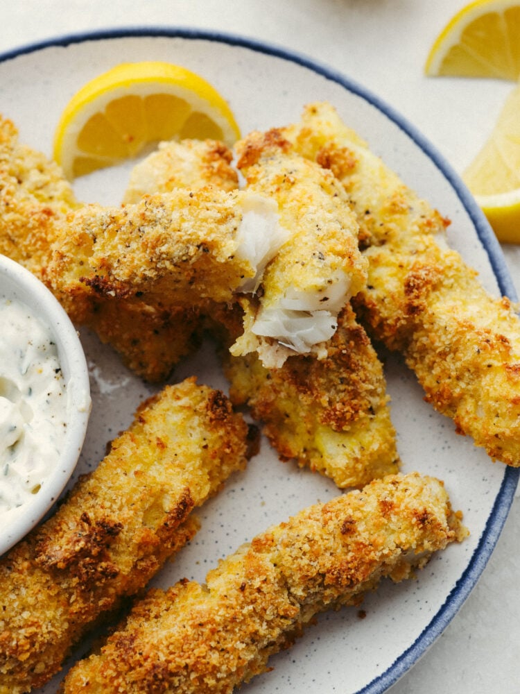 Oven-baked fish sticks are crispy, tender, and SO good! Coated with breadcrumbs and delicious seasoning, these fish sticks are packed with flavor and a recipe that the whole family will enjoy! A plate of fish sticks with one broken open so that you can see the flaky fish inside.