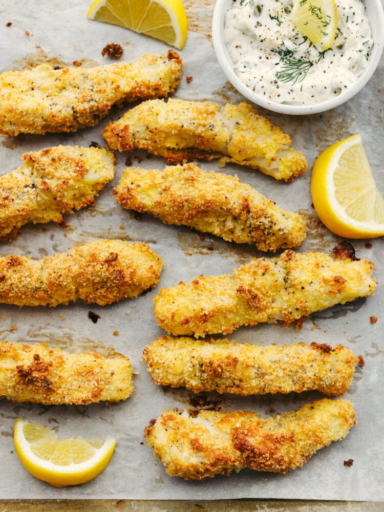 Oven-baked fish sticks are crispy, tender, and SO good! Coated with breadcrumbs and delicious seasoning, these fish sticks are packed with flavor and a recipe that the whole family will enjoy! Baked cod fish sticks on a baking sheet with a slice of lemon and some tarter sauce in a small bowl.