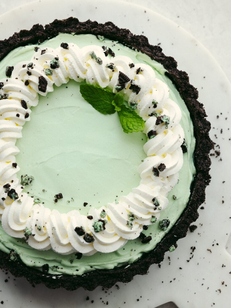 This chilled, minty pie grasshopper pie has a creamy filling sitting on an Oreo cookie crust. The taste and smooth texture are perfect and will have just about anyone coming back for seconds! The top view of the pie that is garnished with cream swirls and crushed andes mints and oreo cookies.