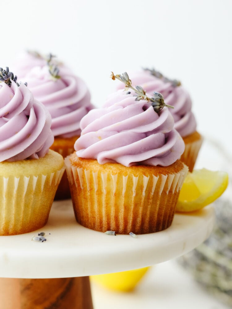 These lovely lavender cupcakes are delicate and delicious. The cake base is lavender flavored and has a lemon buttercream frosting on top, making them perfect for any special occasion! Lavender cupcakes on a cake stand with purple frosting and garnish with lavender buds.