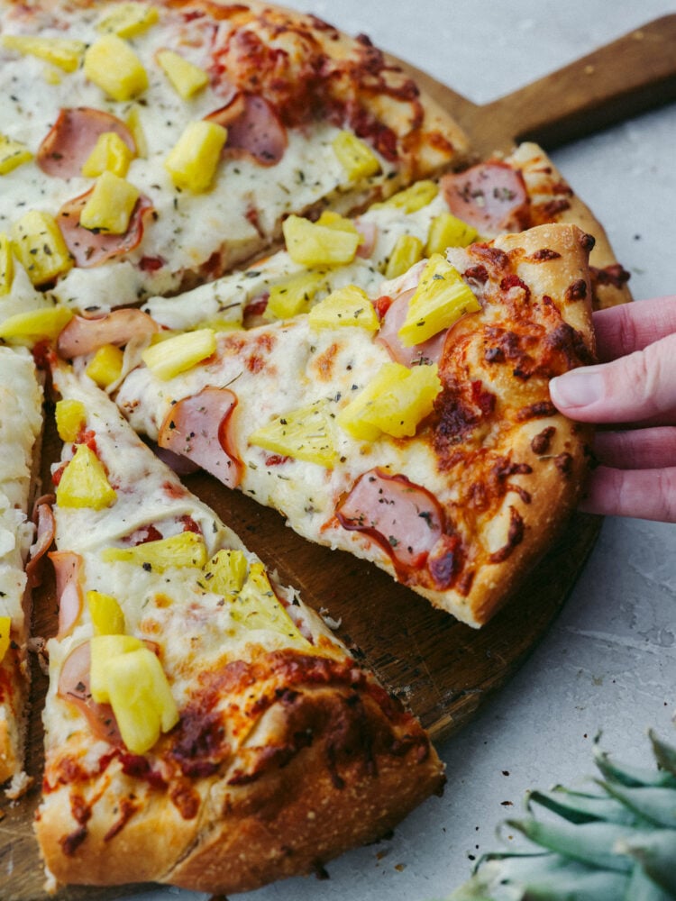 The perfect combination of sweet and savory, Hawaiian pizza is a must-try for your next pizza night! Topped with melty mozzarella, Canadian bacon, pineapple, and Italian herbs, each slice is bursting with flavor! Picking up a slice of pineapple pizza.