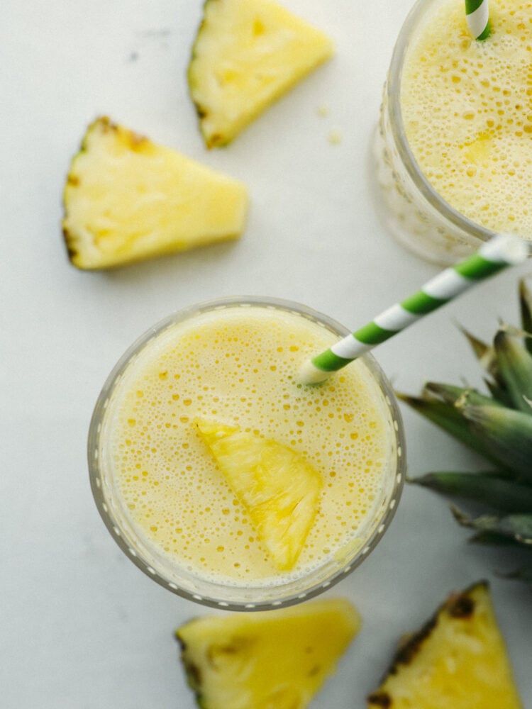 This pineapple smoothie will transport you to a tropical oasis! It has the perfect pineapple flavor combined with orange juice, banana, and some yogurt. If you are looking for the perfect summertime treat, this is it! Top view of a pineapple smoothie in a slass with a chunk of pineapple o top and a green and white straw.