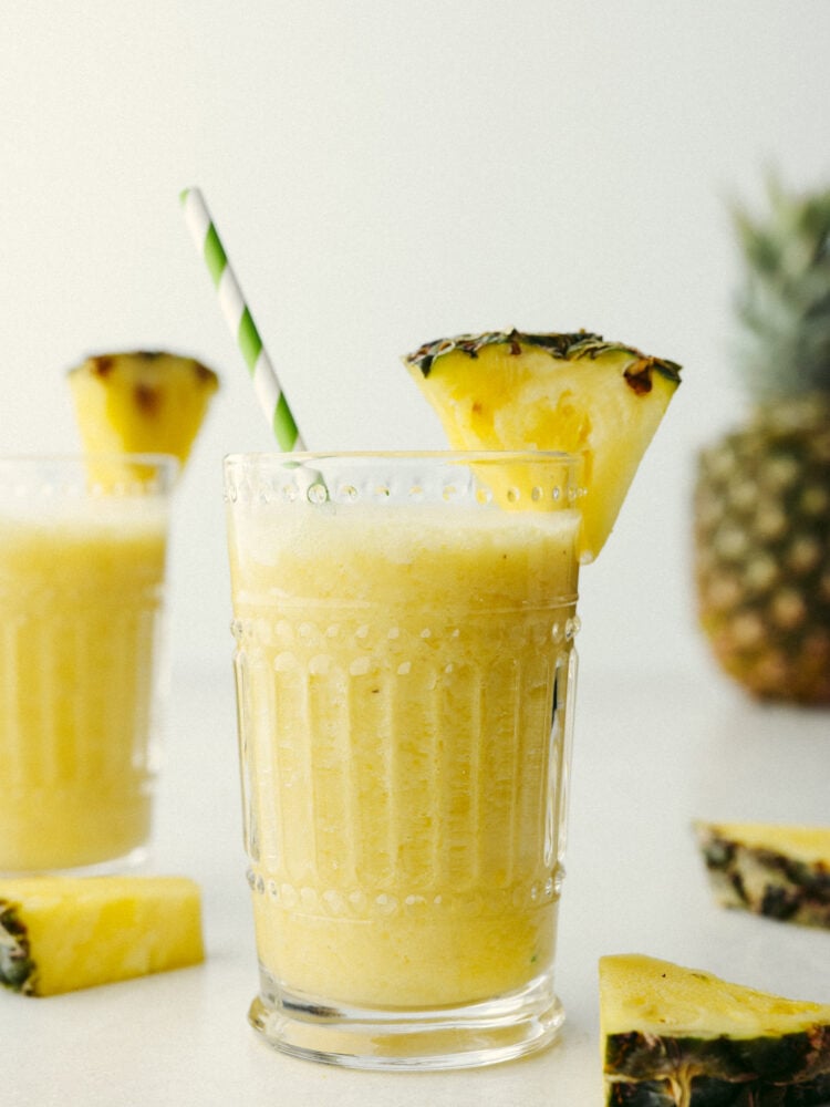This pineapple smoothie will transport you to a tropical oasis! It has the perfect pineapple flavor combined with orange juice, banana, and some yogurt. If you are looking for the perfect summertime treat, this is it! A pineapple smoothie in a glass with a green and white straw and a slice of pineapple on the glass.