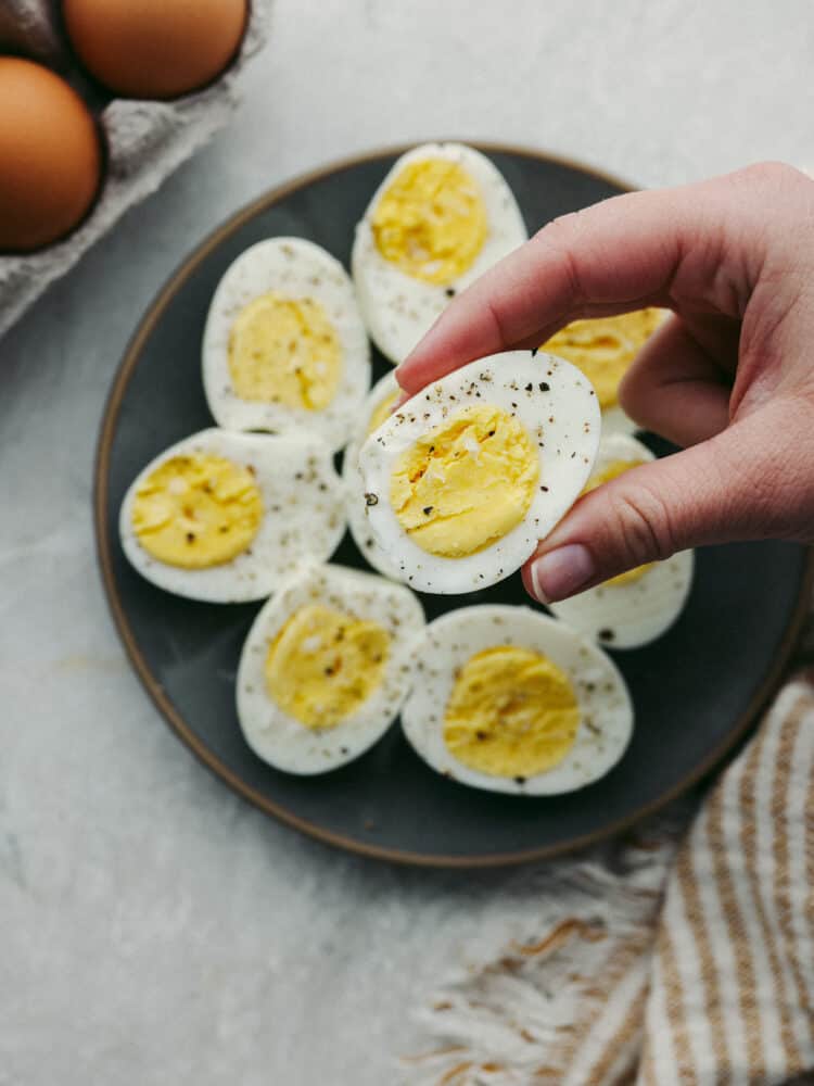 These perfectly cooked air fryer hard-boiled eggs will blow your mind! Convenient, quick, and easy to peel every time, they will be your go-to for some quick protein for any meal! Someone holding the half of a sliced hard boiled egg above a plate filled with hard boiled eggs, sliced and ready to eat.