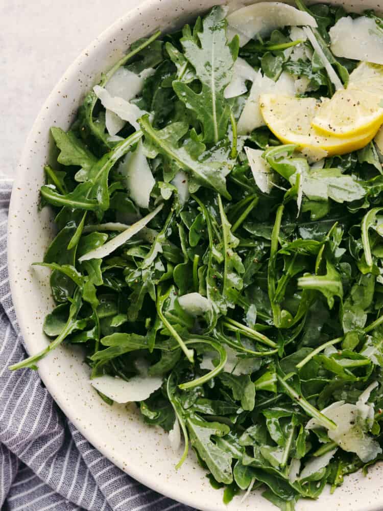 This classic and simple arugula salad will elevate any meal! With only a few ingredients, it comes together quickly, and the flavors of the arugula, Parmesan Reggiano, and lemon vinaigrette really shine together. Arugula salad, garnished with lemon, in a white stoneware bowl.