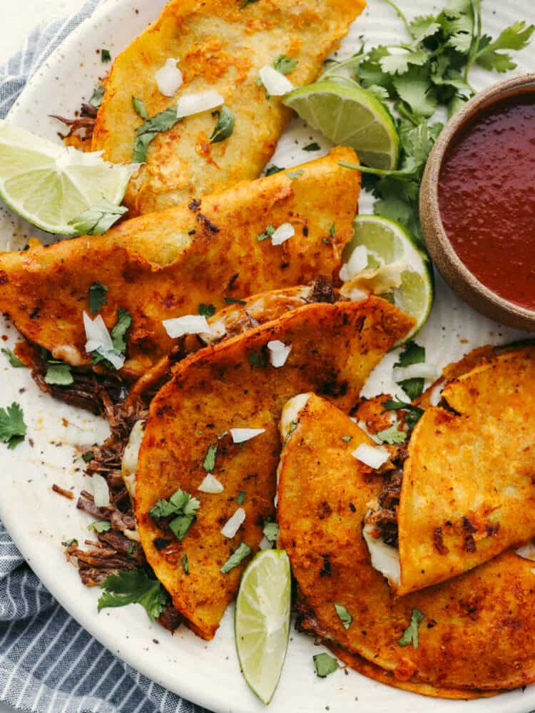 These Birria tacos are packed full of flavor! They are filled with beef and Mexican flavorings that are so delicious that you will definitely need to come to this meal hungry because you will for sure be asking for seconds! A close up of a platter of birria tacos and sauce.